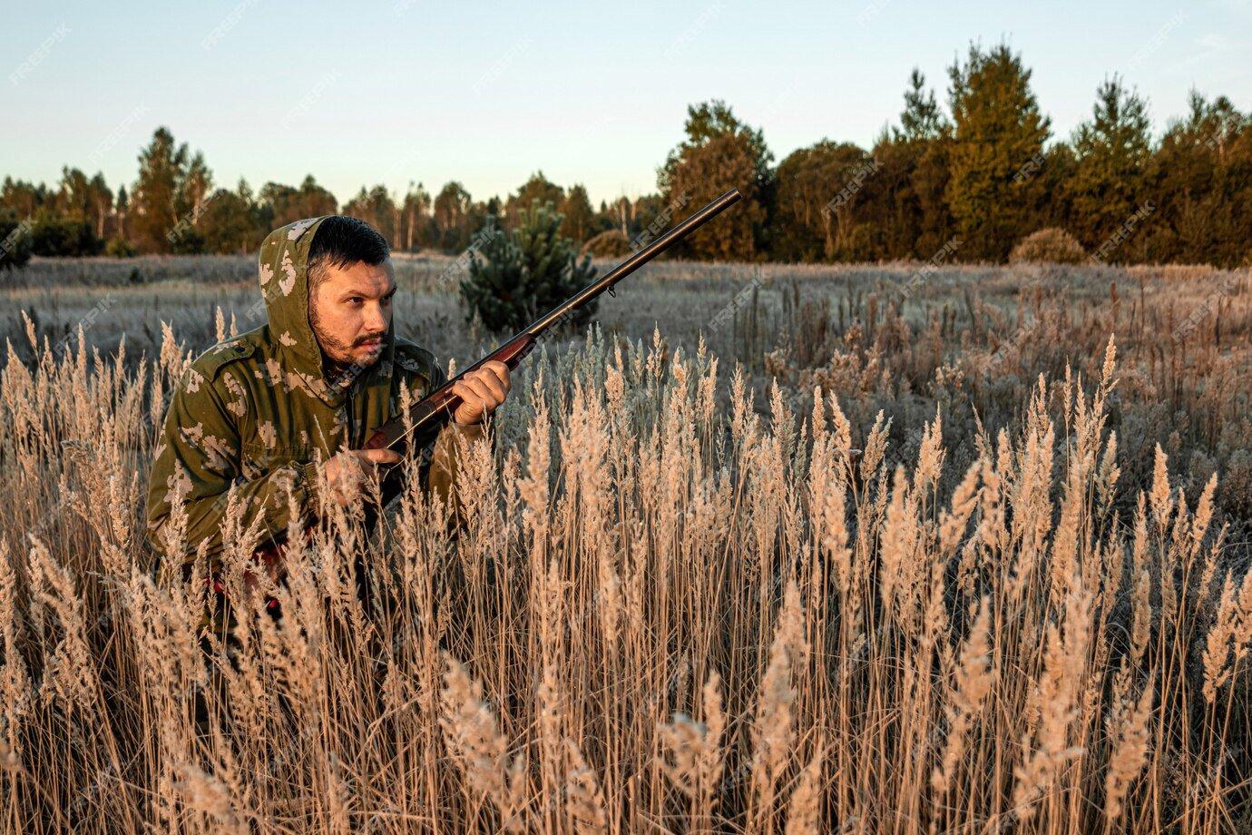 Hunter with a gun is sitting in the grass waiting for prey